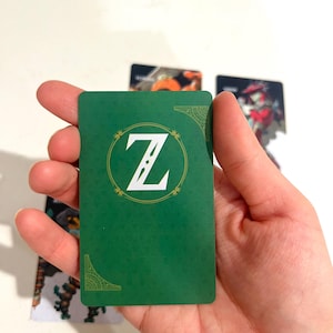 May include: A rectangular playing card with a solid green background featuring a white letter Z centered within a circular gold-toned border. The card includes decorative gold-toned corner accents and a subtle textured pattern across the surface.