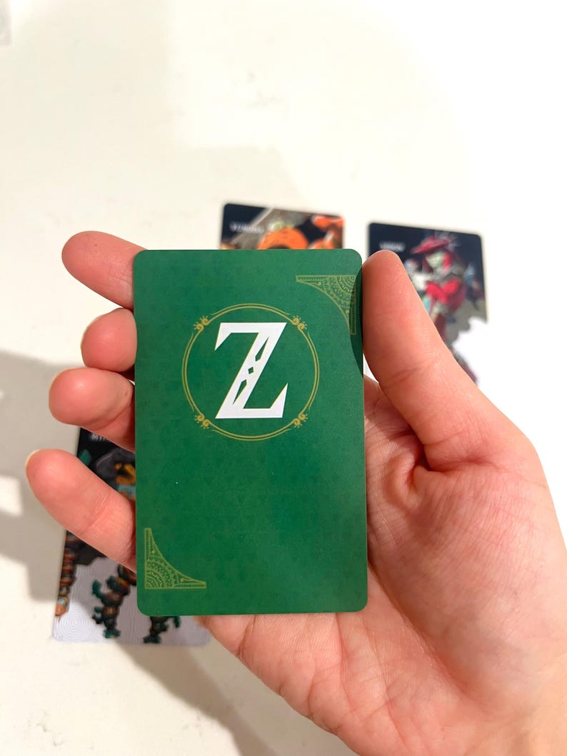 May include: A rectangular playing card with a solid green background featuring a white letter Z centered within a circular gold-toned border. The card includes decorative gold-toned corner accents and a subtle textured pattern across the surface.