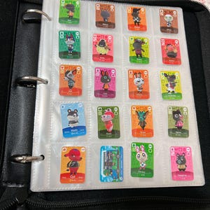 May include: A black three-ring binder containing a clear plastic sheet with nineteen rectangular Animal Crossing trading cards. Each card features a unique character illustration, colourful borders, and text identifying the character name and card number.