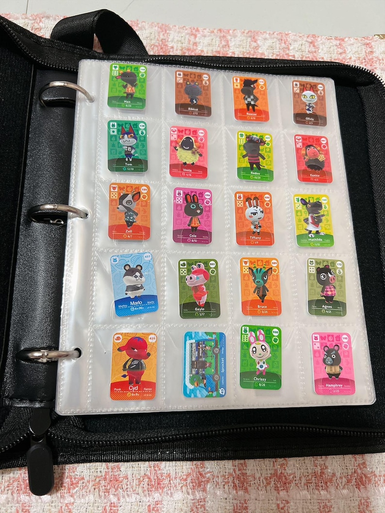 May include: A black three-ring binder containing a clear plastic sheet with nineteen rectangular Animal Crossing trading cards. Each card features a unique character illustration, colourful borders, and text identifying the character name and card number.