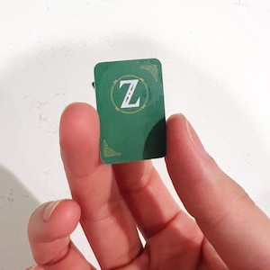 May include: A small rectangular green token featuring a white letter Z centered within a circular gold-toned border. The token has rounded corners and shows subtle decorative gold-toned corner accents on the top right and bottom left edges.