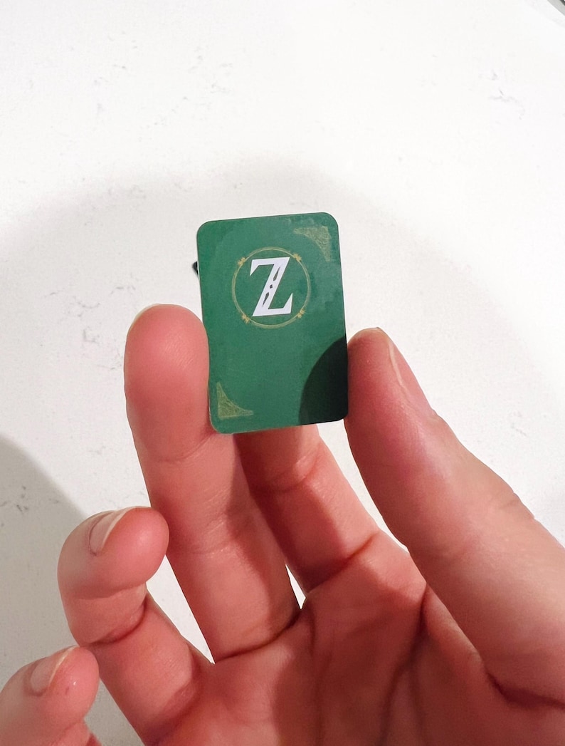 May include: A small rectangular green token featuring a white letter Z centered within a circular gold-toned border. The token has rounded corners and shows subtle decorative gold-toned corner accents on the top right and bottom left edges.