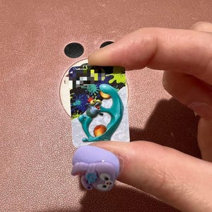 May include: A light purple ring with a cartoon bunny design. The ring is on a finger, with a colorful sticker in the background. The sticker has a teal, orange, and blue design.