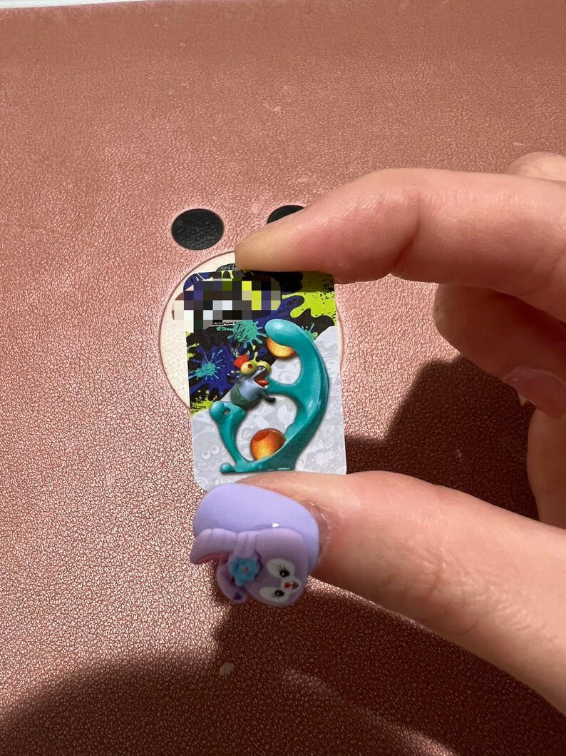 May include: A light purple ring with a cartoon bunny design. The ring is on a finger, with a colorful sticker in the background. The sticker has a teal, orange, and blue design.