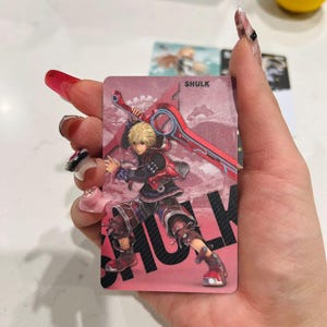 Puede incluir: Una tarjeta rosa con una ilustraci&oacute;n de personaje del videojuego Xenoblade Chronicles. El personaje, Shulk, se representa con cabello rubio y un atuendo rojo y negro, sosteniendo una gran espada roja. El nombre "SHULK" est&aacute; impreso en letras negras grandes.