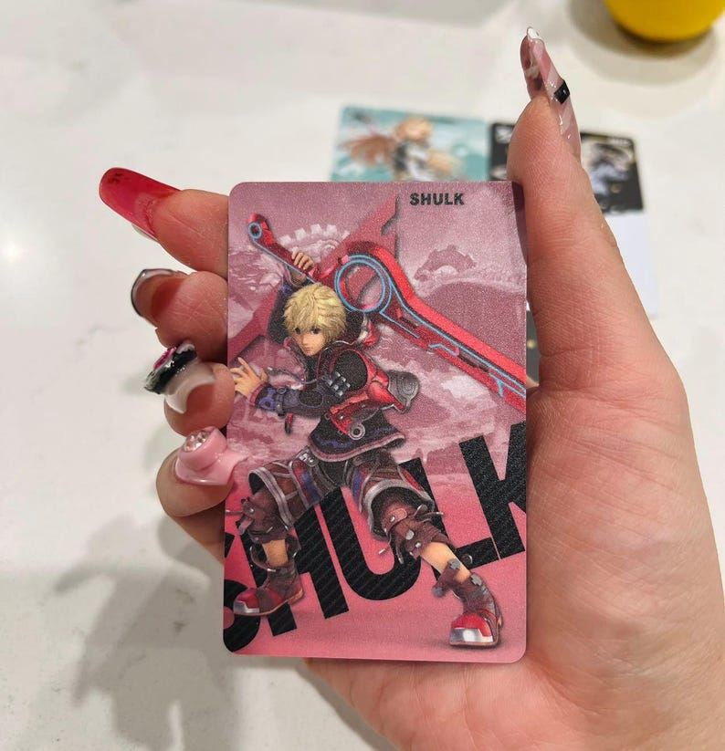 Puede incluir: Una tarjeta rosa con una ilustraci&oacute;n de personaje del videojuego Xenoblade Chronicles. El personaje, Shulk, se representa con cabello rubio y un atuendo rojo y negro, sosteniendo una gran espada roja. El nombre "SHULK" est&aacute; impreso en letras negras grandes.