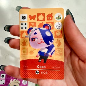 May include: A rectangular trading card featuring a cartoon character named Cece with purple fur, blue hair, and a blue shirt. The card has an orange background with various icons, a red bottom section with the name Cece, and the number 6/28 printed below.