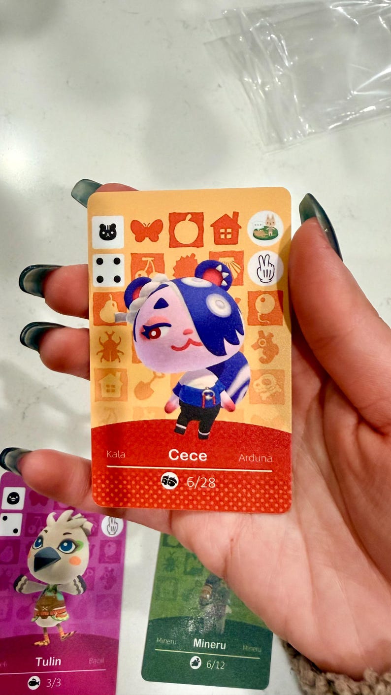 May include: A rectangular trading card featuring a cartoon character named Cece with purple fur, blue hair, and a blue shirt. The card has an orange background with various icons, a red bottom section with the name Cece, and the number 6/28 printed below.