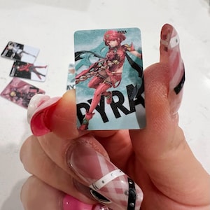 Puede incluir: Una tarjeta rectangular con una ilustraci&oacute;n de personaje en rojo y blanco, y el nombre "PYRA" en negro. Se ven otras tarjetas similares en el fondo. La tarjeta se sostiene en una mano con u&ntilde;as largas y decoradas.