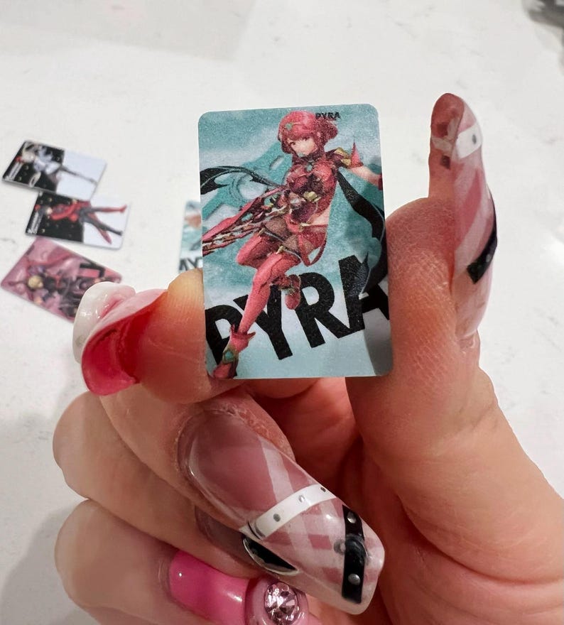 Puede incluir: Una tarjeta rectangular con una ilustraci&oacute;n de personaje en rojo y blanco, y el nombre "PYRA" en negro. Se ven otras tarjetas similares en el fondo. La tarjeta se sostiene en una mano con u&ntilde;as largas y decoradas.