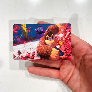 May include: A rectangular card featuring Donkey Kong in a dynamic pose, set against a vibrant background of red and blue hues. The artwork showcases the character's distinctive fur and facial features, with a touch of action-oriented design.