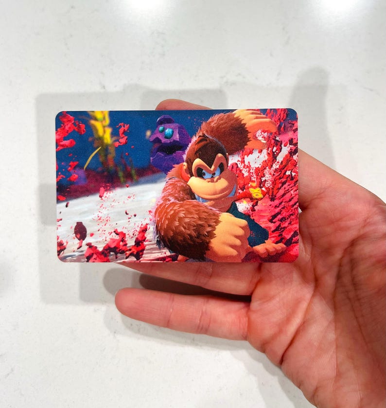 May include: A rectangular card featuring Donkey Kong in a dynamic pose, set against a vibrant background of red and blue hues. The artwork showcases the character's distinctive fur and facial features, with a touch of action-oriented design.