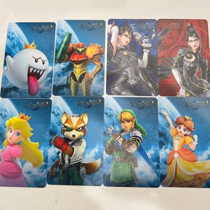 K&ouml;nnte beinhalten: Ein Set aus acht rechteckigen Sammelkarten mit verschiedenen Videospielcharakteren wie King Boo, Samus Aran, Bayonetta, Prinzessin Peach, Fox McCloud, Link und Prinzessin Daisy, alle vor einem blauen Himmelshintergrund mit dem Bayonetta 2-Logo.