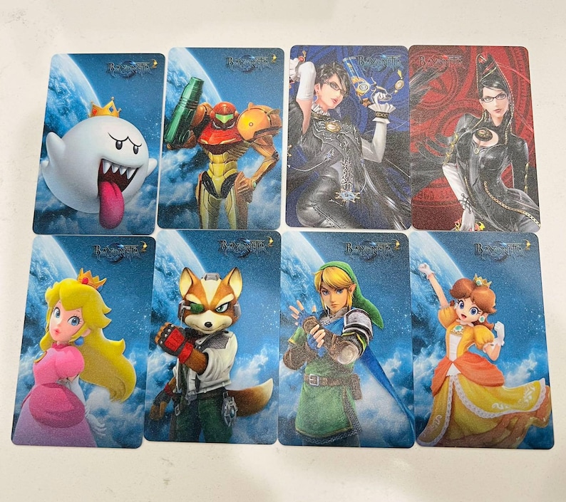 K&ouml;nnte beinhalten: Ein Set aus acht rechteckigen Sammelkarten mit verschiedenen Videospielcharakteren wie King Boo, Samus Aran, Bayonetta, Prinzessin Peach, Fox McCloud, Link und Prinzessin Daisy, alle vor einem blauen Himmelshintergrund mit dem Bayonetta 2-Logo.
