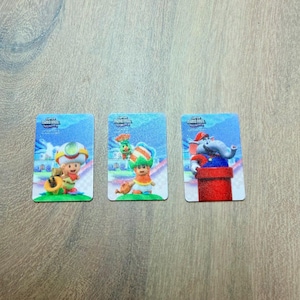 Puede incluir: Tres tarjetas rectangulares con personajes de Super Mario Bros. Wonder. Cada tarjeta tiene un fondo azul con una ilustraci&oacute;n de personaje. Los personajes incluyen Toad, un personaje con un sombrero verde y naranja y un personaje de elefante.