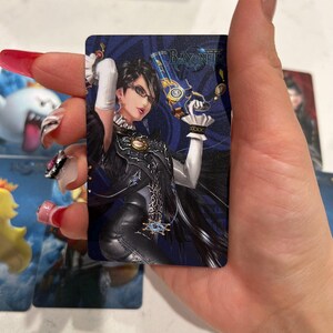 K&ouml;nnte beinhalten: Eine rechteckige Sammelkarte mit einer dunkelhaarigen Figur mit Brille, die ein schwarzes Outfit und wei&szlig;e Handschuhe tr&auml;gt und eine gold-blaue Handfeuerwaffe h&auml;lt. Die Karte zeigt den Schriftzug Bayonetta in der oberen rechten Ecke auf blauem Grund.