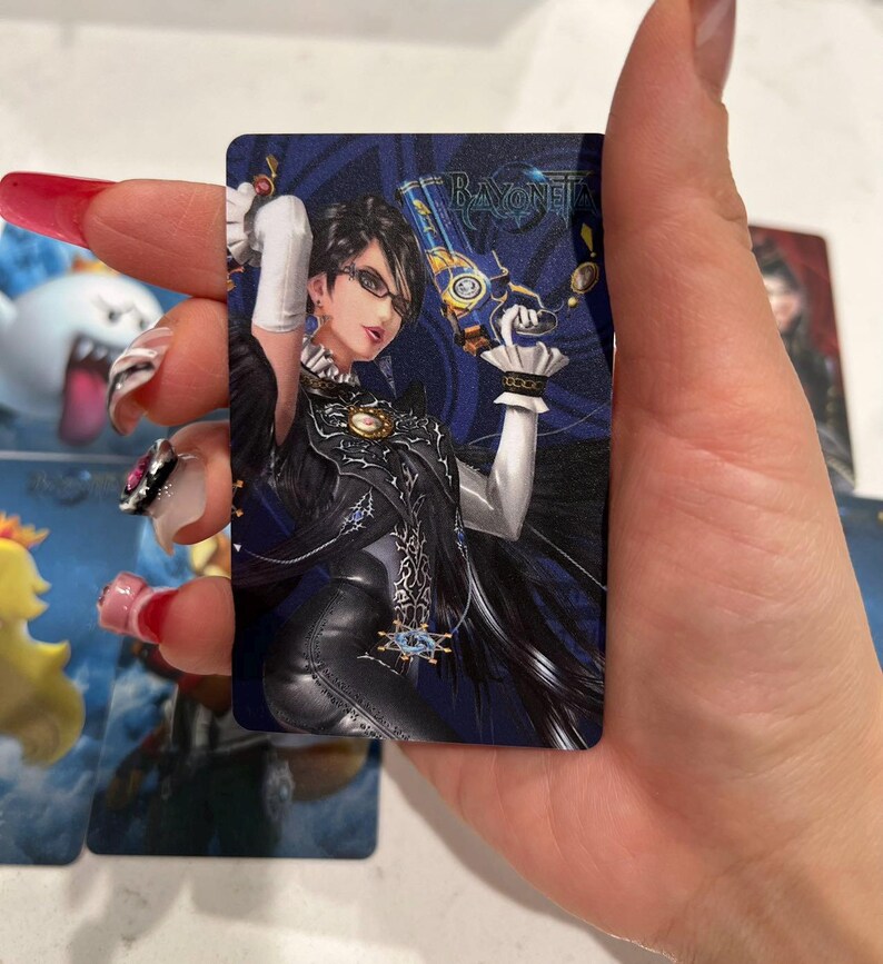 K&ouml;nnte beinhalten: Eine rechteckige Sammelkarte mit einer dunkelhaarigen Figur mit Brille, die ein schwarzes Outfit und wei&szlig;e Handschuhe tr&auml;gt und eine gold-blaue Handfeuerwaffe h&auml;lt. Die Karte zeigt den Schriftzug Bayonetta in der oberen rechten Ecke auf blauem Grund.