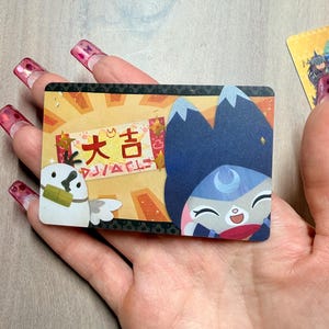 May include: A rectangular card featuring cartoon illustrations and Japanese text. The card has a light yellow background with a cartoon owl and a blue-haired character. The Japanese text reads "大吉" and "DJ/ACLE".