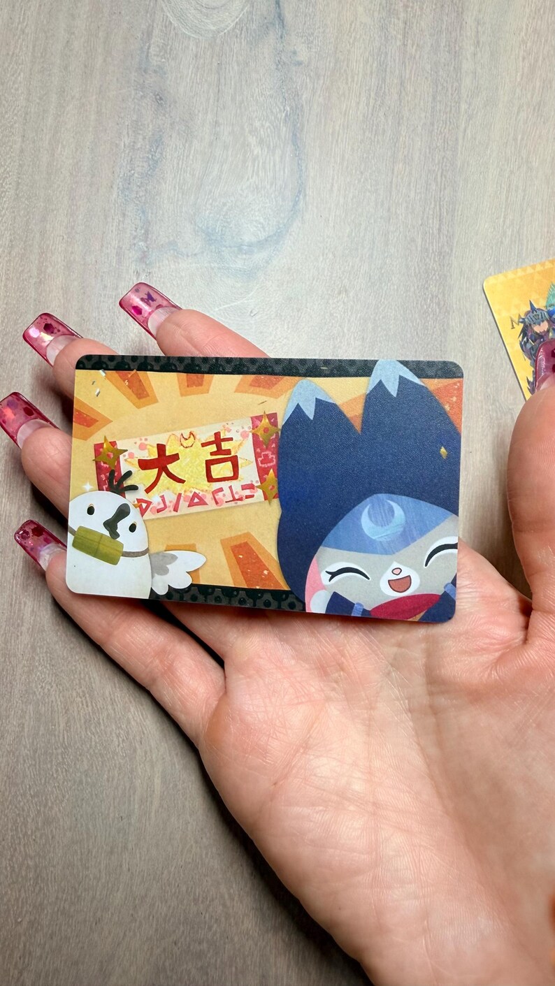 May include: A rectangular card featuring cartoon illustrations and Japanese text. The card has a light yellow background with a cartoon owl and a blue-haired character. The Japanese text reads "大吉" and "DJ/ACLE".