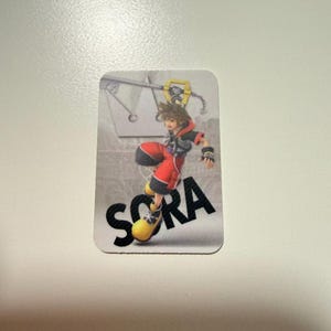 May include: A rectangular sticker featuring the character Sora from Kingdom Hearts in a dynamic pose, wearing a black and red outfit with large yellow shoes. The background is a light gray gradient with a crown icon and the name SORA in bold black text.