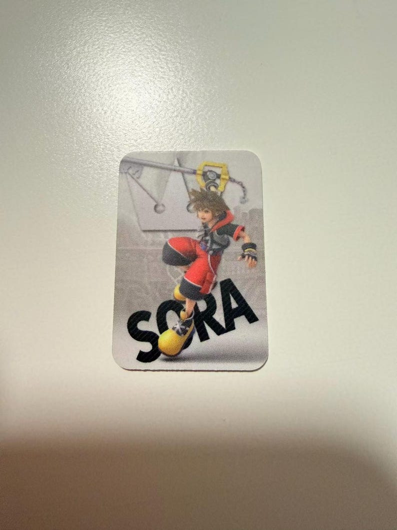 May include: A rectangular sticker featuring the character Sora from Kingdom Hearts in a dynamic pose, wearing a black and red outfit with large yellow shoes. The background is a light gray gradient with a crown icon and the name SORA in bold black text.