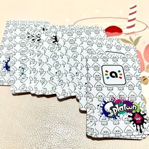 May include: A deck of white playing cards featuring a repeating pattern of cartoon squid figures. The cards have colorful ink splatters and the "Splatoon" logo. The back of the cards has a logo with a colorful "a" inside a square.