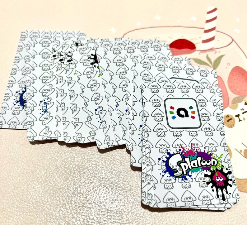 May include: A deck of white playing cards featuring a repeating pattern of cartoon squid figures. The cards have colorful ink splatters and the "Splatoon" logo. The back of the cards has a logo with a colorful "a" inside a square.