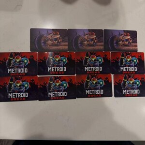 May include: A collection of rectangular cards featuring artwork from the video game Metroid Dread. The cards display images of a character in a suit and a vehicle, with the game's title prominently displayed.