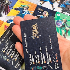 May include: A rectangular black game card featuring The Legend of Zelda Breath of the Wild logo, text listing Link Ocarina of Time, Cap of Time, Tunic of Time, Trousers of Time, and Biggoron's Sword, surrounded by various colorful character cards.