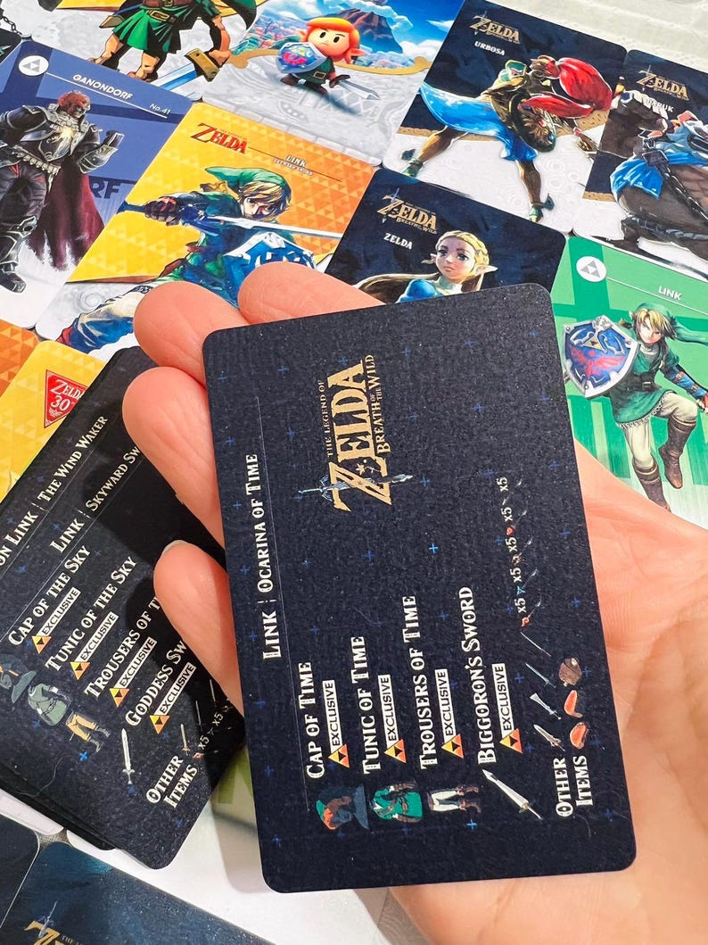 May include: A rectangular black game card featuring The Legend of Zelda Breath of the Wild logo, text listing Link Ocarina of Time, Cap of Time, Tunic of Time, Trousers of Time, and Biggoron's Sword, surrounded by various colorful character cards.