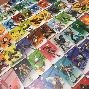 May include: A collection of rectangular trading cards featuring various video game characters, each with a unique illustration and name printed on the card. The cards are arranged in a grid pattern, displaying colorful artwork of characters like Kirby, Samus, Yoshi, and Falco.