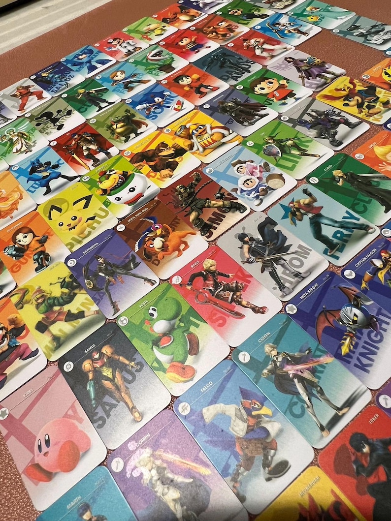 May include: A collection of rectangular trading cards featuring various video game characters, each with a unique illustration and name printed on the card. The cards are arranged in a grid pattern, displaying colorful artwork of characters like Kirby, Samus, Yoshi, and Falco.