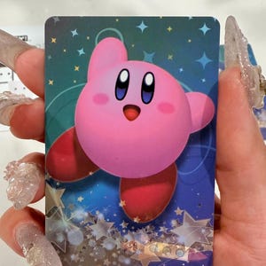 Op de afbeelding: Een rechthoekige verzamelkaart met het ronde, roze Kirby-personage met grote blauwe ogen en rode voeten. De achtergrond is een diepblauw verloop gevuld met lichtblauwe en gouden sterren van verschillende groottes, wat een sprankelend effect cre&euml;ert.