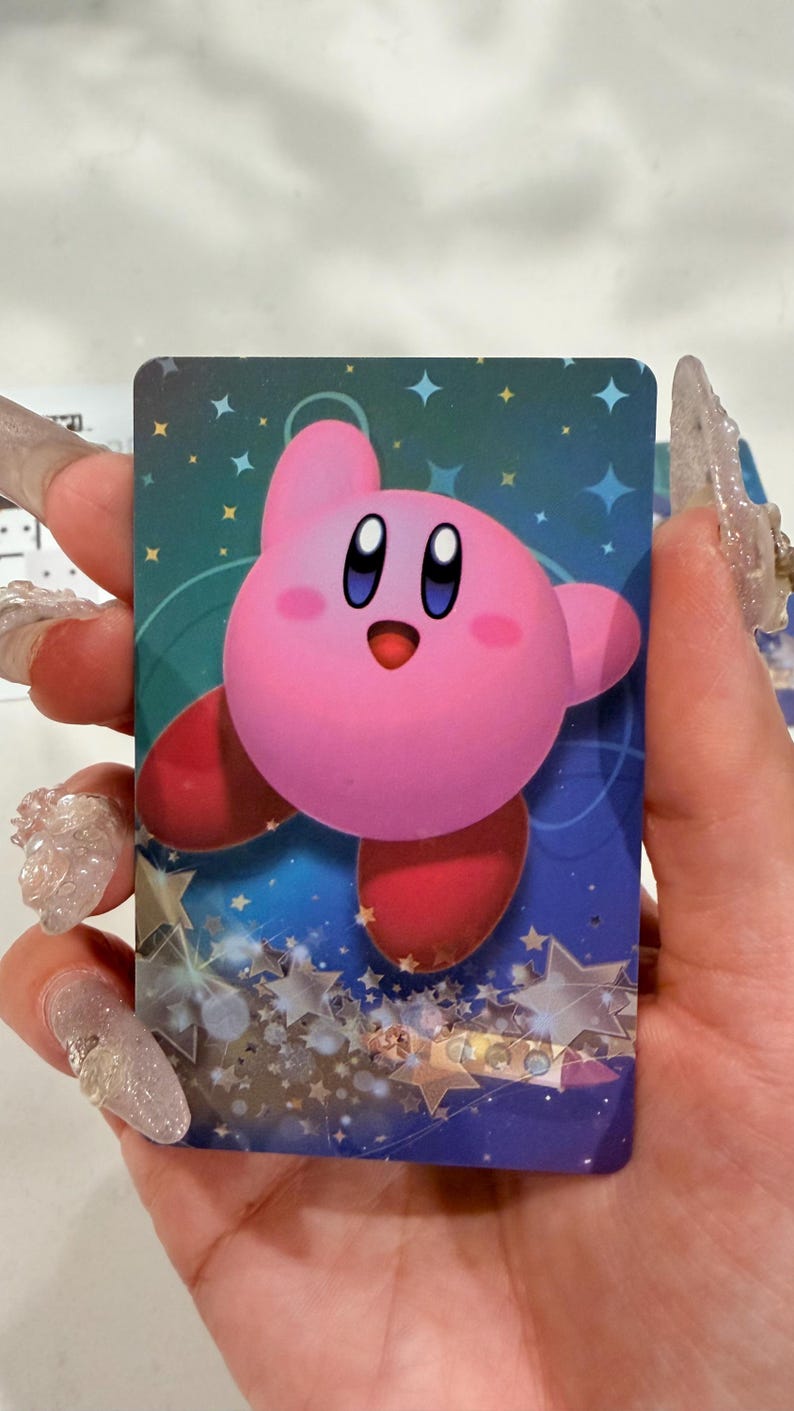 Op de afbeelding: Een rechthoekige verzamelkaart met het ronde, roze Kirby-personage met grote blauwe ogen en rode voeten. De achtergrond is een diepblauw verloop gevuld met lichtblauwe en gouden sterren van verschillende groottes, wat een sprankelend effect cre&euml;ert.