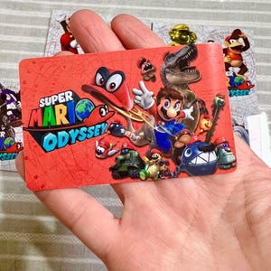Op de afbeelding: Een rechthoekige Super Mario Odyssey spelkaart met een felrode achtergrond en een kaartpatroon. Het ontwerp toont Mario, Cappy, een T-Rex en diverse vijanden uit het spel. Het Super Mario Odyssey logo staat linksboven afgedrukt.