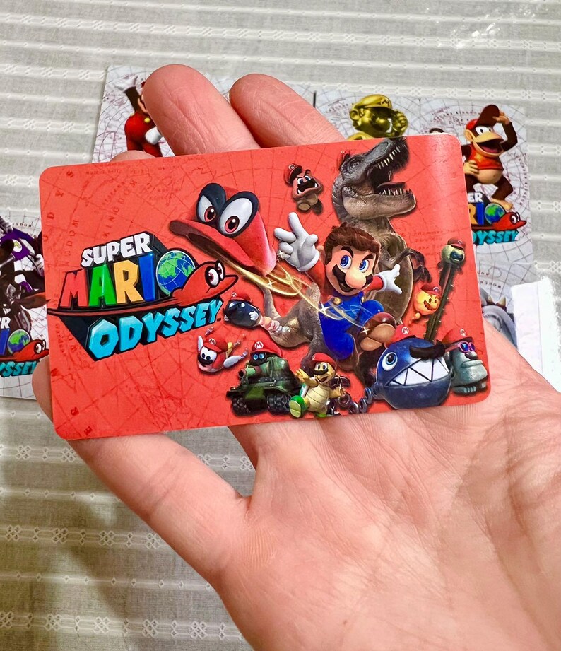 Op de afbeelding: Een rechthoekige Super Mario Odyssey spelkaart met een felrode achtergrond en een kaartpatroon. Het ontwerp toont Mario, Cappy, een T-Rex en diverse vijanden uit het spel. Het Super Mario Odyssey logo staat linksboven afgedrukt.