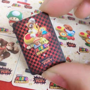 以下が含まれることがあります： スーパーマリオ3Dワールドのキャラクターが描かれた長方形のステッカー。ステッカーは赤と黒のチェック柄の背景です。キャラクターにはマリオ、猫、その他のゲーム要素が含まれています。「Super Mario 3D World Bowser's Fury」の文字も表示されています。