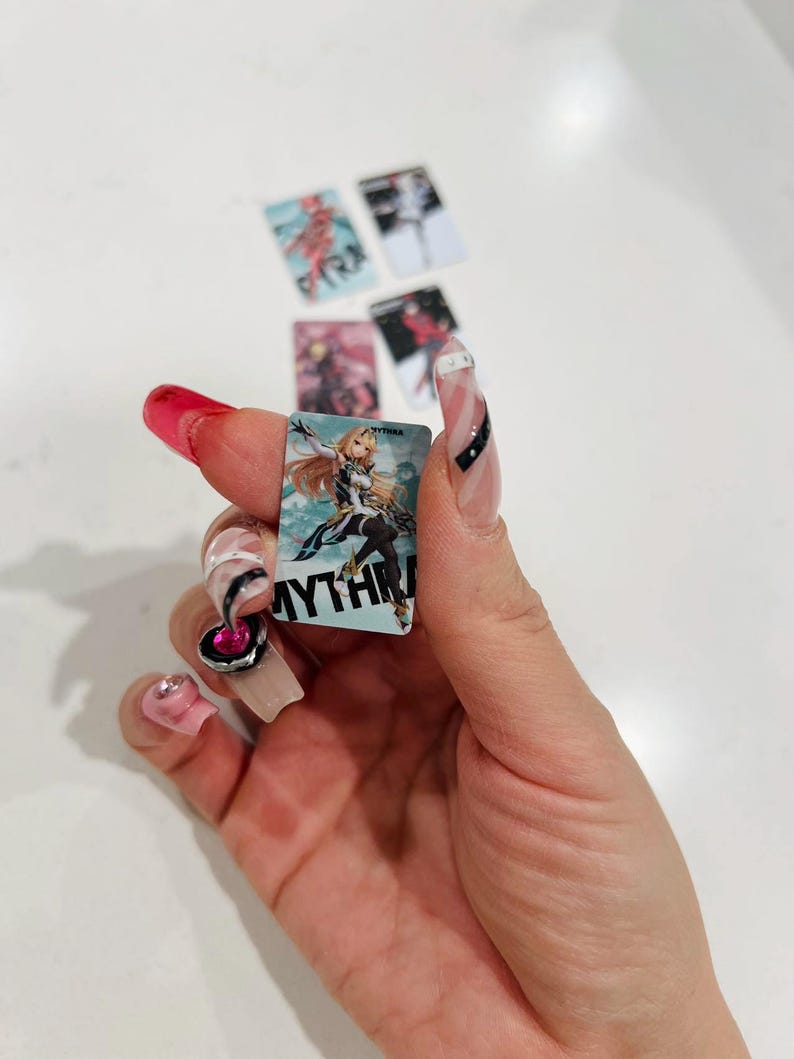 Puede incluir: Peque&ntilde;as tarjetas rectangulares con ilustraciones de estilo anime. Una tarjeta muestra un personaje rubio con la palabra "MYTHRA" impresa. Otras tarjetas muestran diferentes personajes. Las tarjetas se sostienen en una mano con u&ntilde;as largas y decoradas.