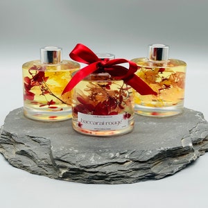 May include: Three glass bottles filled with a clear liquid and dried flowers. The bottles have silver caps and red ribbon bows. The bottles are sitting on a grey slate surface. The bottle in the centre has a label that reads "baccarai rouge".
