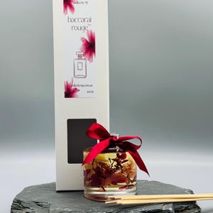 May include: A clear glass diffuser bottle with a red ribbon and dried flowers inside. The bottle is sitting on a black slate surface with wooden diffuser sticks. The bottle is labelled "Candles by MJ Baccarat Rouge 200ml candlesbymj.com.au"