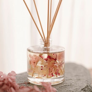 May include: A clear glass bottle diffuser with dried pink and white flowers inside. The bottle has wooden sticks sticking out of the top. The bottle is sitting on a grey stone.