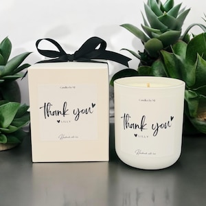 May include: A white candle and a matching box tied with a black ribbon. Both the candle and box feature a label with the words "thank you" in script, the name "Lilly", and the phrase "Handmade with love". The candle is in a white glass container.