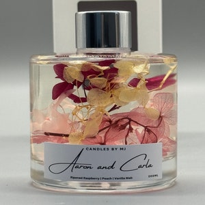 May include: A clear glass diffuser bottle with a silver lid. The bottle contains dried pink and white flowers. The label reads "Candles by MJ Aaron and Carla Ripened Raspberry | Peach | Vanilla Malt 200ML".