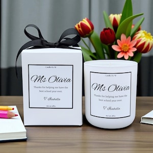 May include: A white candle and gift box set tied with a black ribbon. Both items have a black framed label with the text "Ms Olivia" and a thank you message. A bouquet of flowers is in the background.