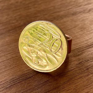 May include: A gold-colored coin ring featuring a 50-cent coin. The coin is mounted on a brown band. The ring is set against a wooden surface. The coin's details are visible, including the number "50".