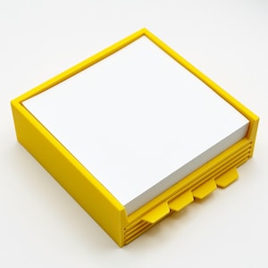 May include: A bright yellow square notepad holder with a stack of white paper. The holder has a frame and tabs on the side. The notepad is a simple and functional office supply.
