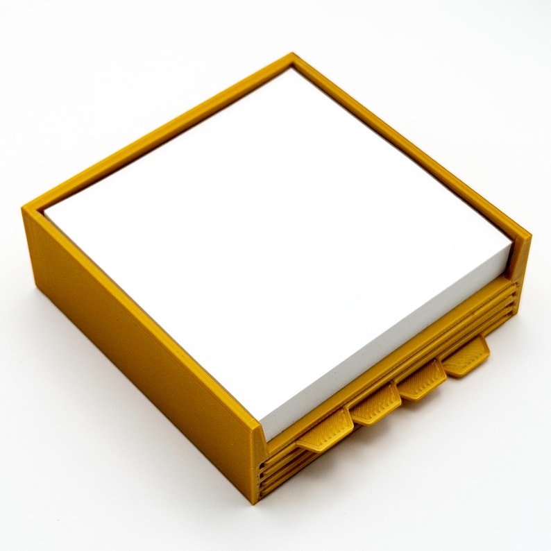 May include: Mustard yellow square notepad holder with a stack of white notepaper. The holder has a rectangular frame and four built-in tabs. The tabs are used to separate the notepaper. The notepad holder is made of plastic.