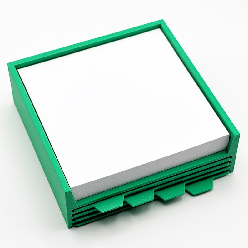 May include: A green square notepad holder with a stack of white paper. The holder has a rectangular frame and a series of tabs on the bottom edge. The design is simple and modern, suitable for office or home use.