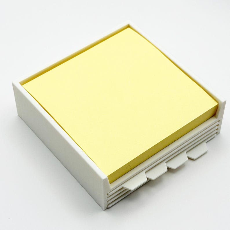 May include: A white, square sticky note holder with a stack of yellow sticky notes. The holder has a stepped design on the front with small tabs. The holder is made of plastic and is designed to hold a pad of sticky notes.