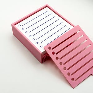 May include: A pink desk organizer with a stack of lined white notepaper and a pink divider. The notepaper has blue lines and circles. The divider has slots and holes. The organizer is on a white surface.